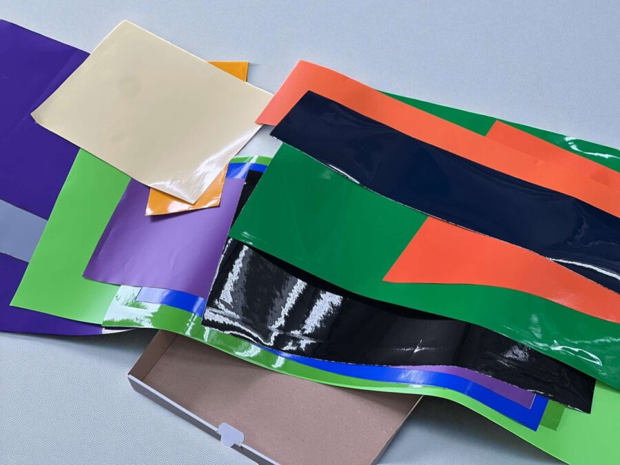 Verschiedenfarbige selbstklebende Folienreste in glänzender Optik liegen auf einem Tisch neben einem offenen Karton.