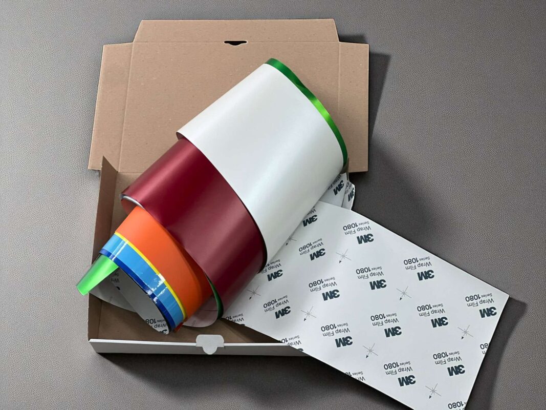 Mehrere bunte Rollen Autofolie in einer offenen Versandverpackung, darunter auch Folien von 3M mit sichtbarem Rückseitenaufdruck.