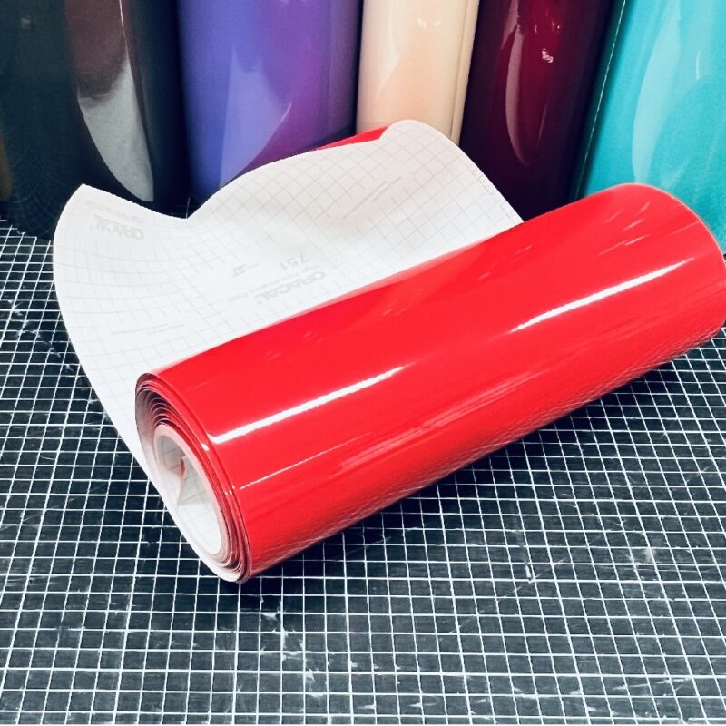 ORACAL 324 Blutrot glänzende Plotterfolie mit weißem Trägerpapier auf einer karierten Schneidematte und farbigen Folienrollen im Hintergrund