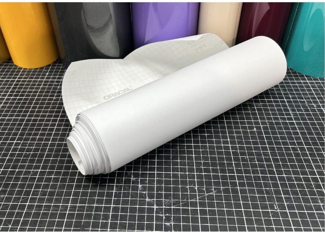 ORACAL 751C-010 Weiß matt als aufgerollte Plotterfolie auf einer karierten Schneidematte, mit weißem Trägerpapier und farbigen Folien im Hintergrund